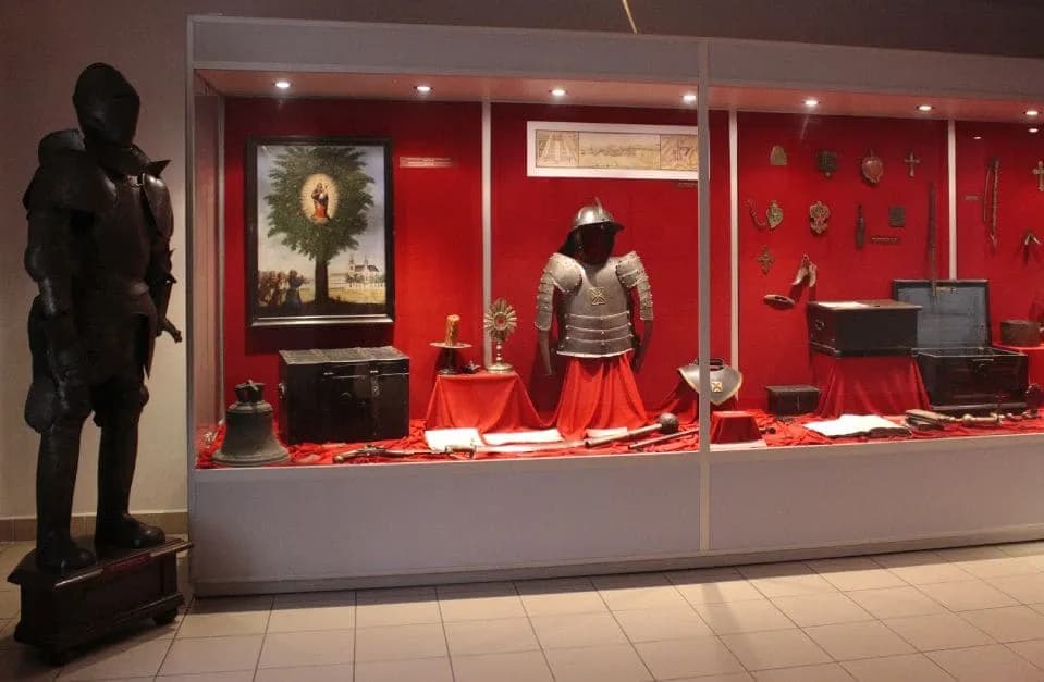 A museum display features a full suit of dark armor on the left, and a long glass case with a red background showcasing another suit of armor, antique weapons, artifacts, and a painting of a tree with a figure inside a halo.