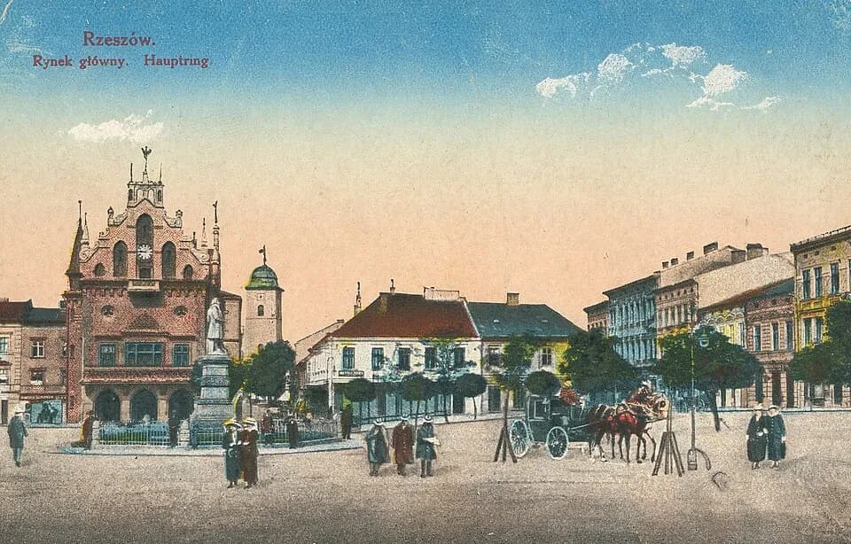 A vintage postcard showing the Rynek Główny market square in Rzeszów, Poland, with a detailed central building, monuments, people, and a horse-drawn carriage in front of other buildings.